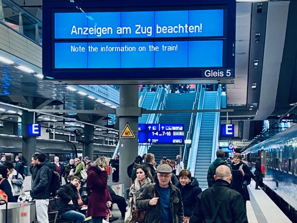 Bahnsteig, wartende Fahrgäste rechts ein Zug. Darüber eine blaue Anzeigetafer auf der auf Deutsch und English steht "Anzeigen am Zug beachten" 