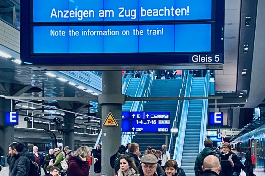 Bahnsteig, wartende Fahrgäste rechts ein Zug. Darüber eine blaue Anzeigetafer auf der auf Deutsch und English steht "Anzeigen am Zug beachten"