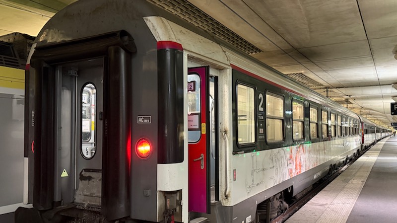 A long passenger train with red and white doors is stopped at an indoor station platform. The train’s lights are on and the platform appears empty.